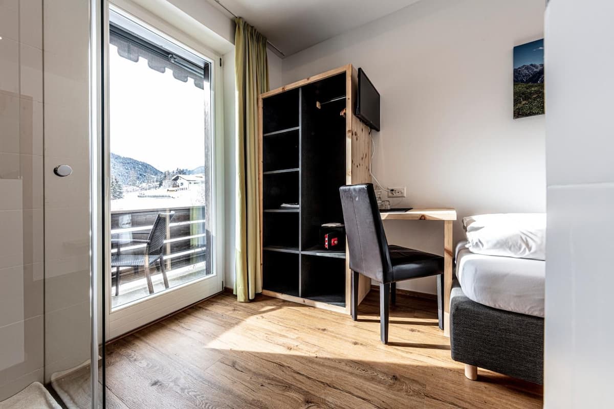 Quarto de hotel com piso de madeira, uma cama, escrivaninha com cadeira, TV fixada na parede e janela com vista para uma paisagem nevada.