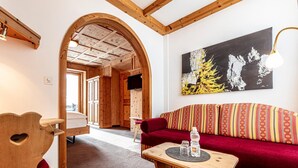 A cozy room with a wooden bench, a red sofa, and a wooden table with a bottle. An archway leads to a hallway with wooden doors and a television. - Hotel Filli (Scuol)