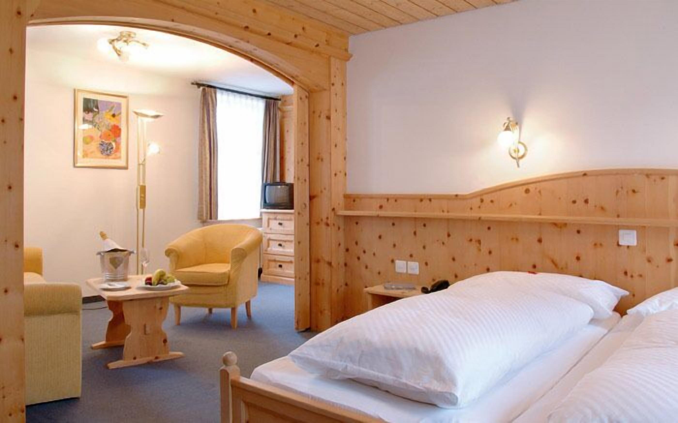 A hotel room with a wooden headboard, a bed with white linens, a chair, a small table with a bottle and fruit, and a television.