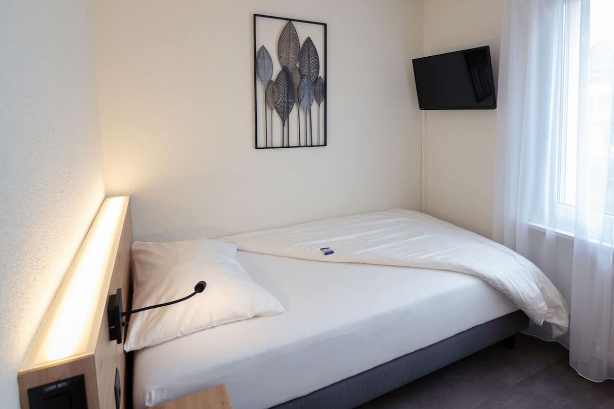 A single bed with white bedding, a bedside table with a lamp, a wall-mounted TV, and a window with sheer curtains.