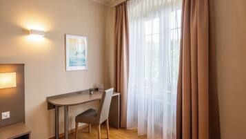 Quarto de hotel com escrivaninha, cadeira e uma janela grande com cortinas leves.