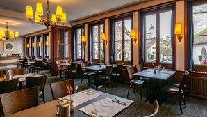 Un restaurante con varias mesas y sillas, amplias ventanas con cortinas y un candelabro.