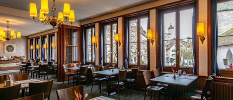 A restaurant with multiple tables and chairs, large windows with curtains, and a chandelier.