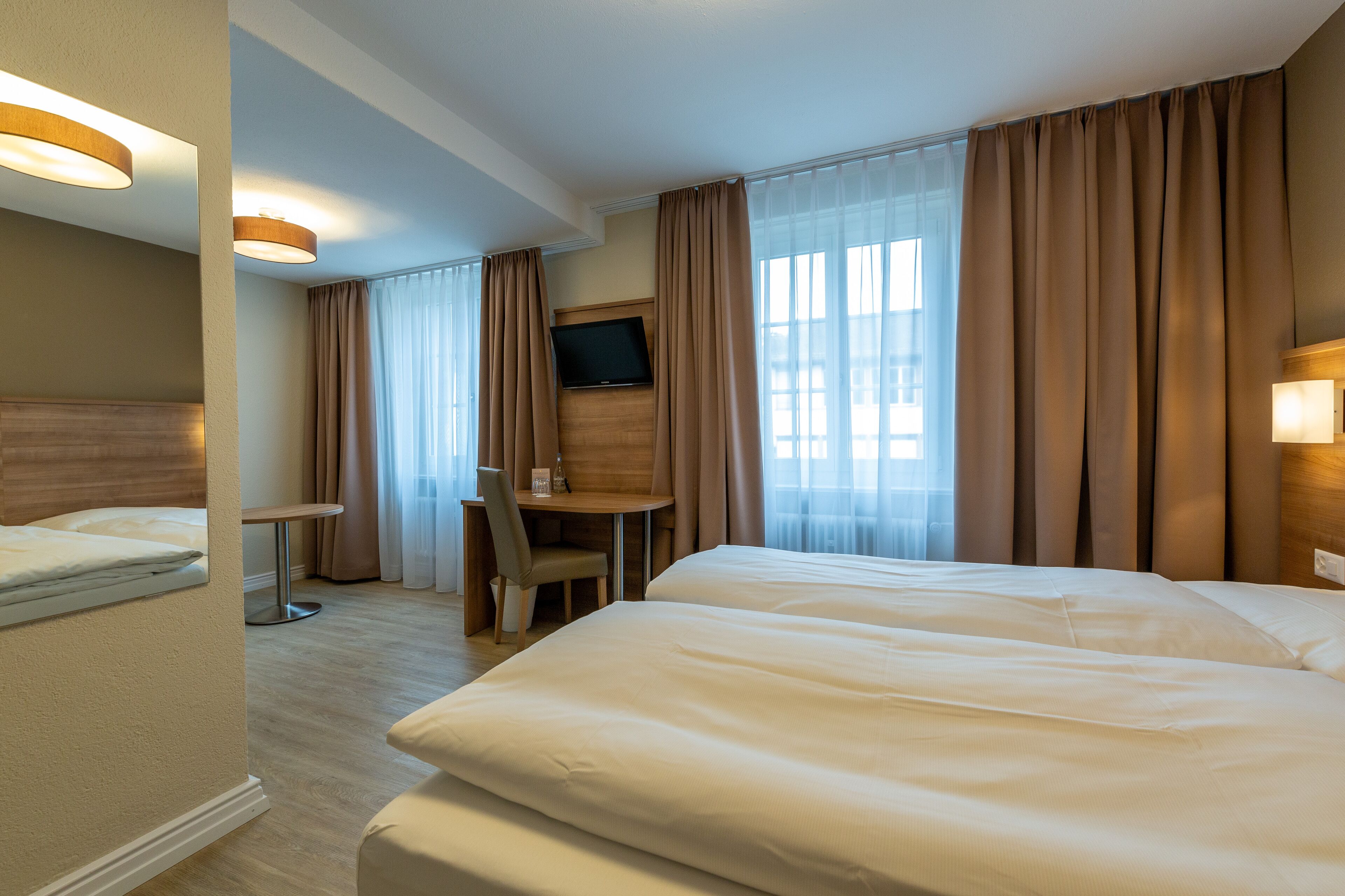 A hotel room with two beds, a desk, a chair, a TV, and large windows with curtains.