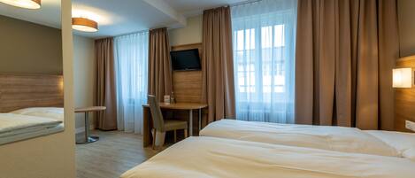 A hotel room with two beds, a desk, a chair, a TV, and large windows with curtains.