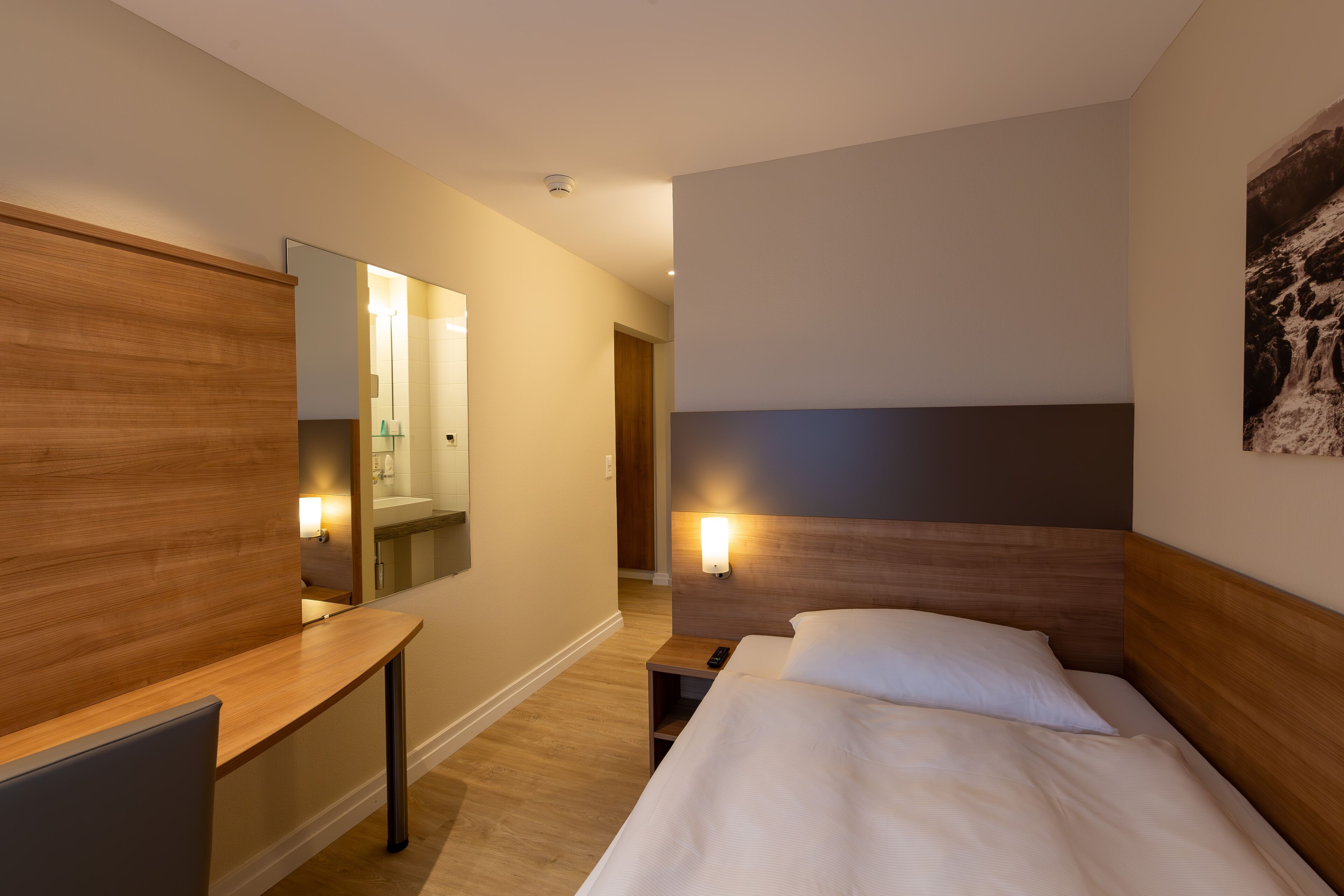 A hotel room with a wooden desk, a bed with white linens, a nightstand with a lamp, and a large mirror on the wall.