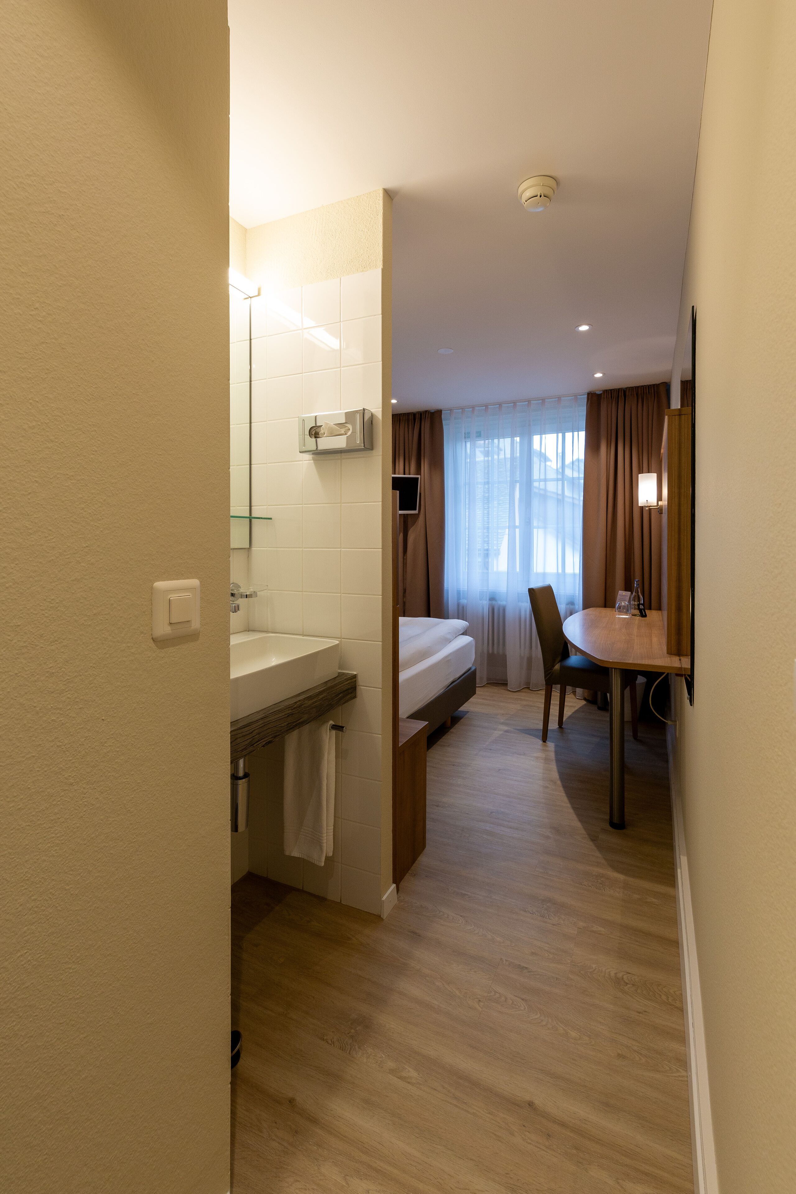 A hotel room with a bed, a bathroom with a sink and mirror, a dining table with chairs, and a window with curtains.