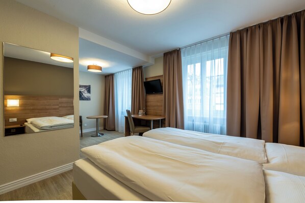 Hotellihuone, jossa on kaksi sänkyä, työpöytä, tuoli, televisio ja suuri ikkuna, jossa on verhot.