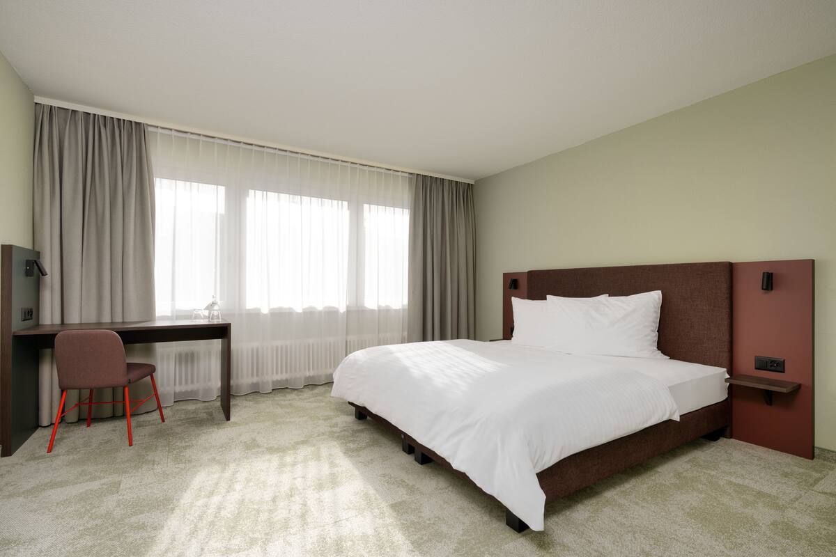 A hotel room with a bed, a desk, a chair, and a window with curtains.