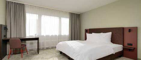 Una habitación de hotel con cama, escritorio, silla y ventana con cortinas.
