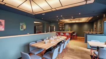 A modern restaurant with a long wooden table, blue chairs, and a bar area.