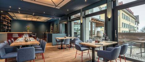 A modern dining area with tables and chairs, a kitchen area, and large windows offering views of buildings.