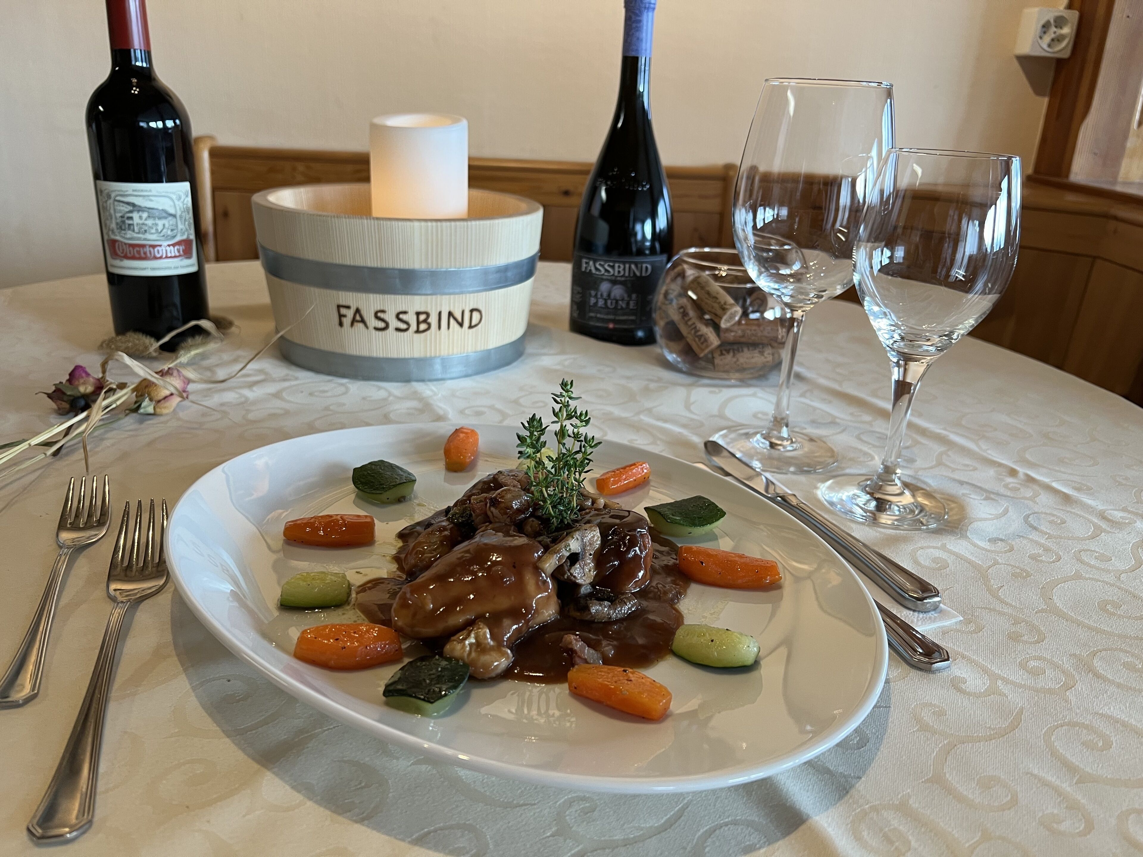 Una mesa de comedor con un plato de comida, botellas de vino, copas y una vela.