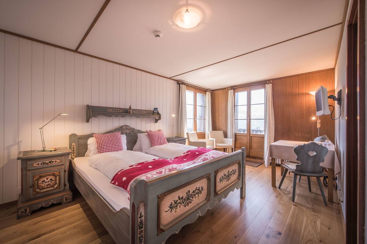 Un dormitorio con cama de madera, mesita de noche, una silla, una mesa y una ventana con cortinas.