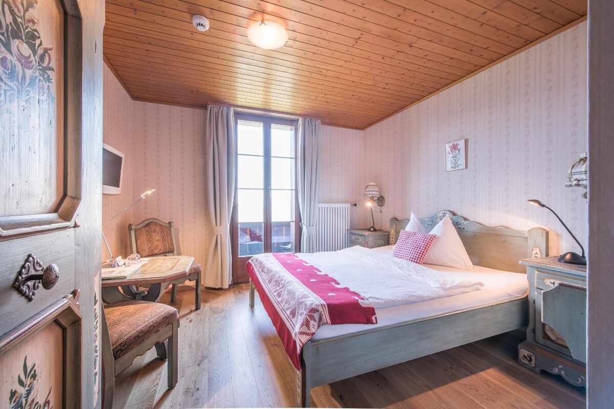 A bedroom with a wooden ceiling, a bed with white bedding, a desk with a chair, and a window with curtains.