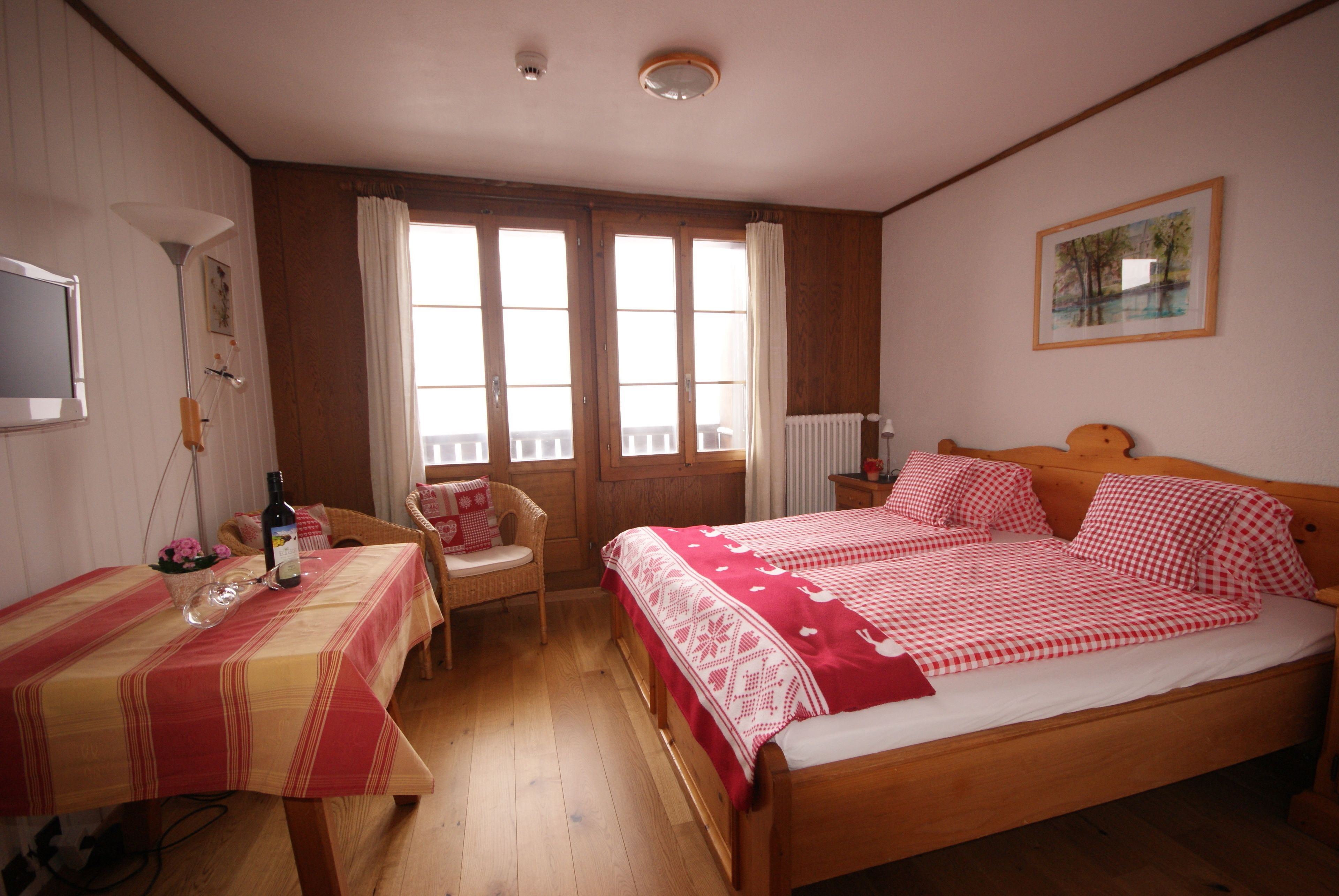 A bedroom with a wooden bed, a dining table with a red and yellow tablecloth, a wicker chair, and a window with curtains.