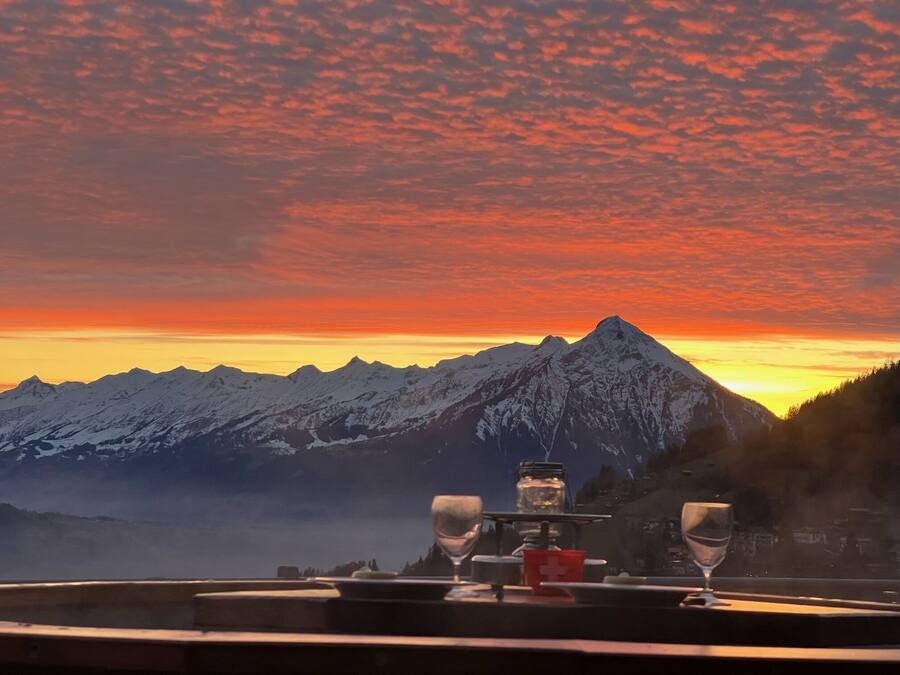 A table with two wine glasses and a small red container, set against a backdrop of snow-capped mountains and a sunset sky.