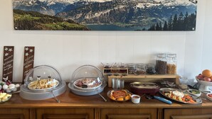 A buffet setup with various food items and condiments, a mountain landscape painting on the wall, and a wooden counter. - Hotel Fassbind Beausite (Beatenberg)