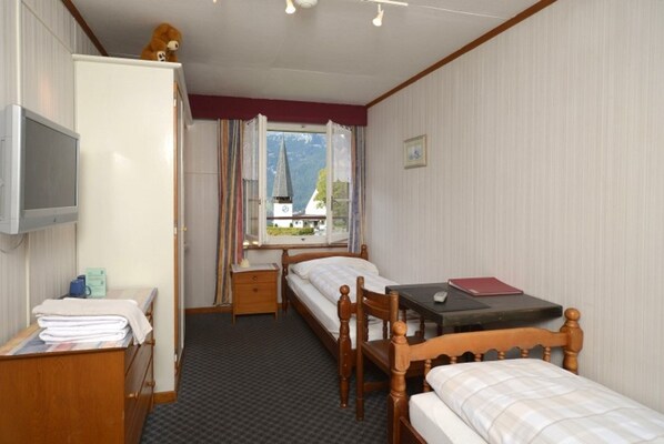 A hotel room with a bed, a desk, a wardrobe, and a window with a view of a mountain and a church steeple. - Swiss Lodge Hotel Bernerhof Wengen (Lauterbrunnen)