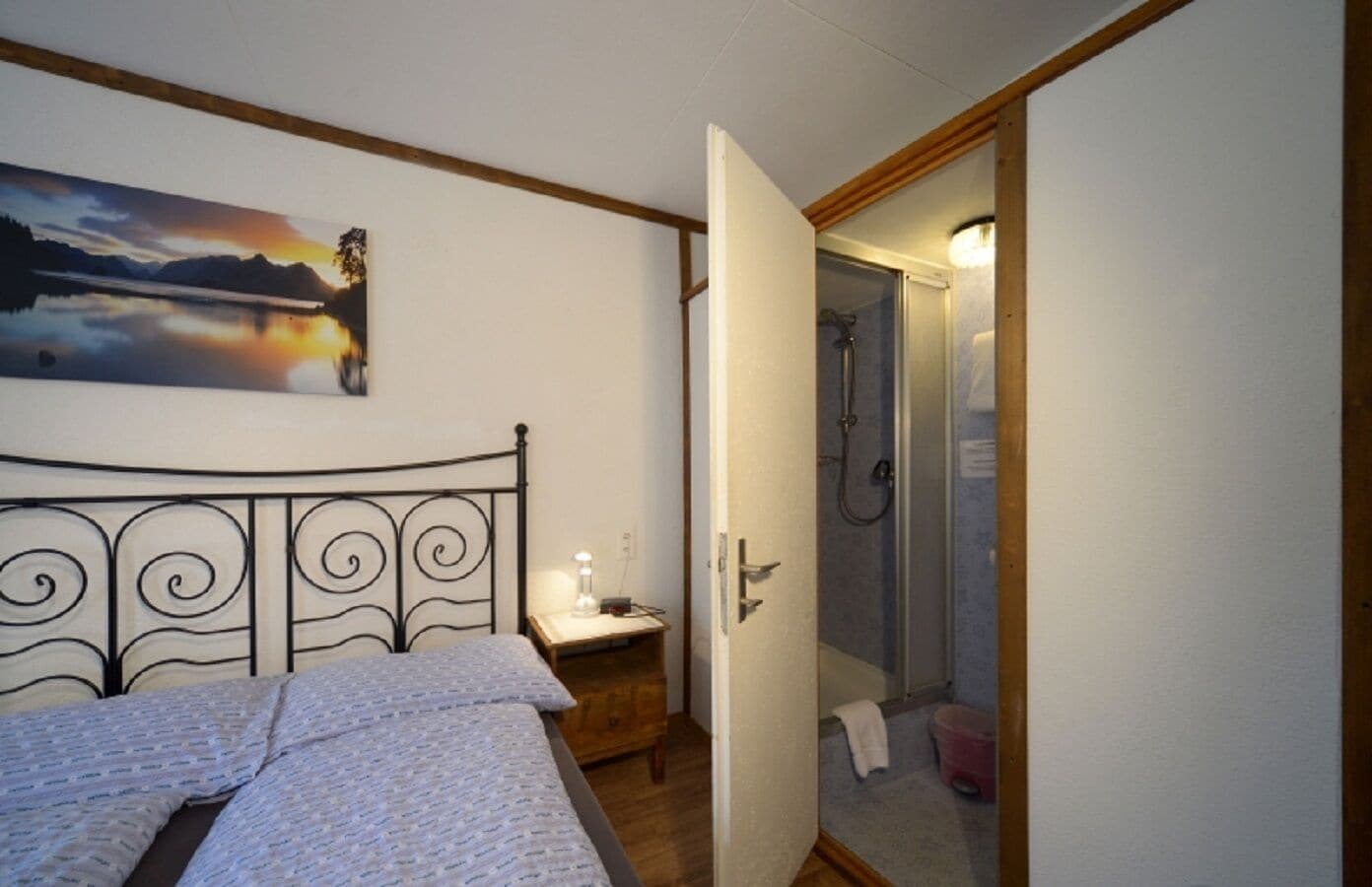A bedroom with a bed, a nightstand, a painting, and an open door leading to a bathroom with a shower.