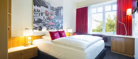 Una habitación de hotel con una cama grande, una mesita de noche, un escritorio, una ventana con cortinas rojas y un televisor montado en la pared.