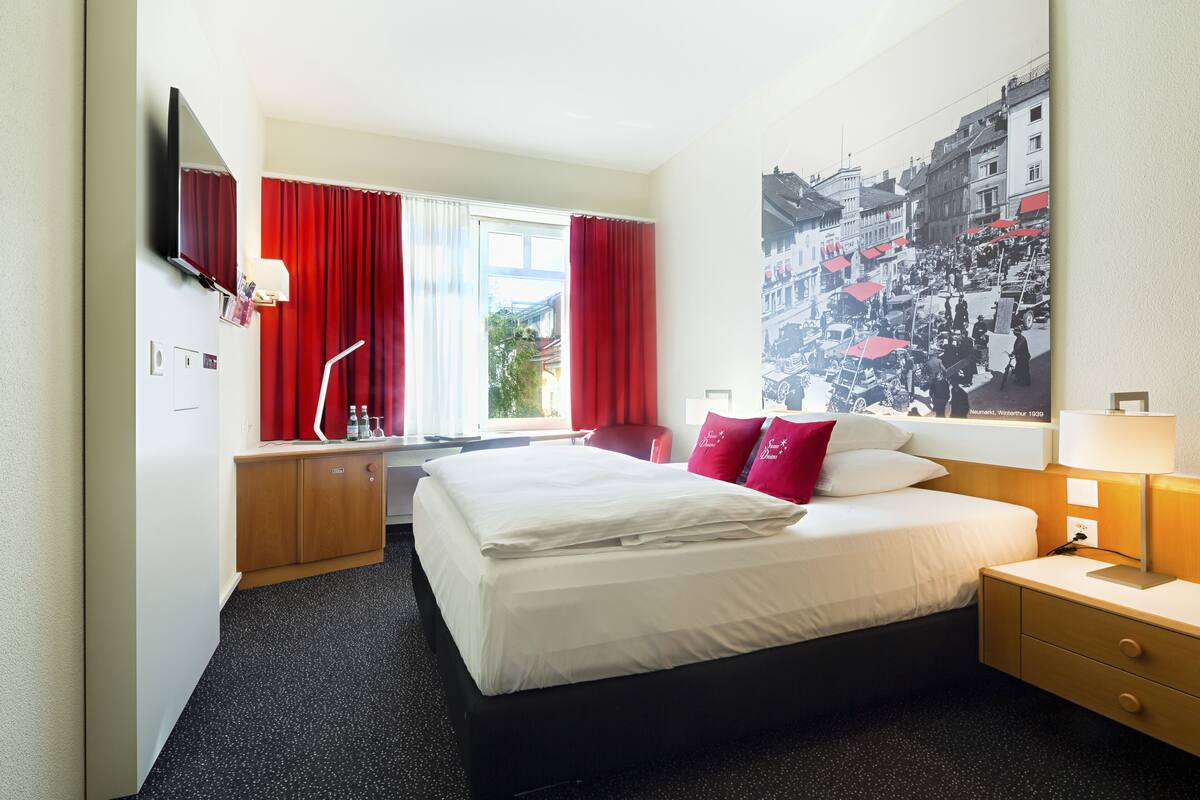 Een hotelkamer met een groot bed, een bureau, een stoel, een televisie en een raam met rode gordijnen.
