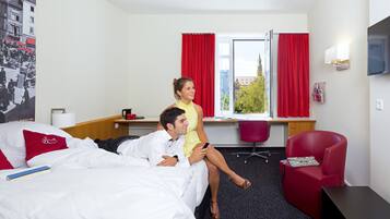 Quarto de hotel com cama, escrivaninha, cadeira e televisão.