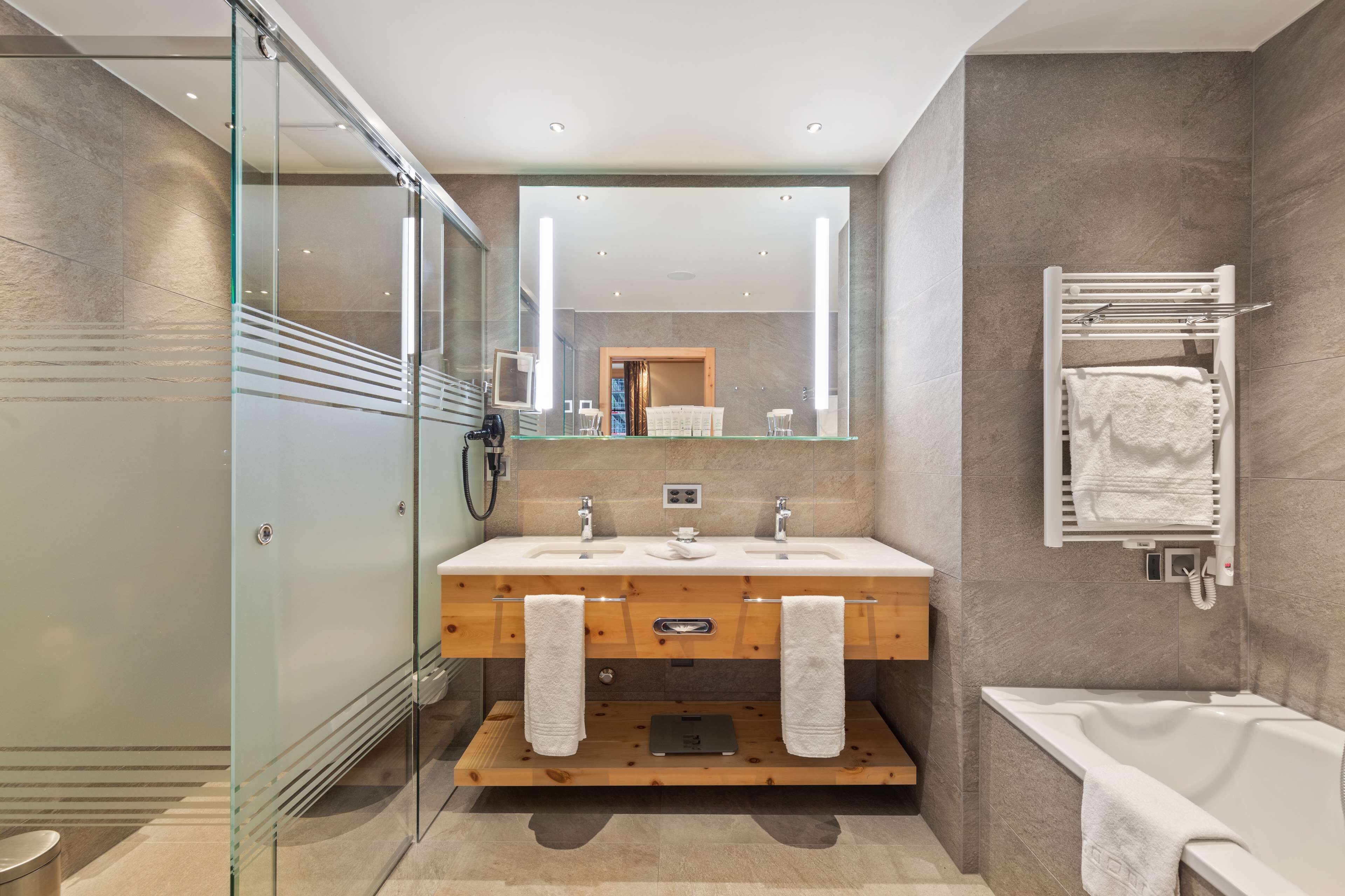Ein modernes Badezimmer mit Duschkabine aus Glas, einem Holzbadezimmerhocker mit zwei Waschbecken, einem großen Spiegel und einer Badewanne.