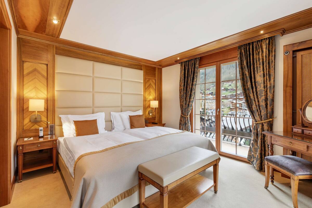 A hotel room with a large bed, wooden headboard, two bedside tables with lamps, a bench, a desk with a chair, and a view of a balcony with a railing.