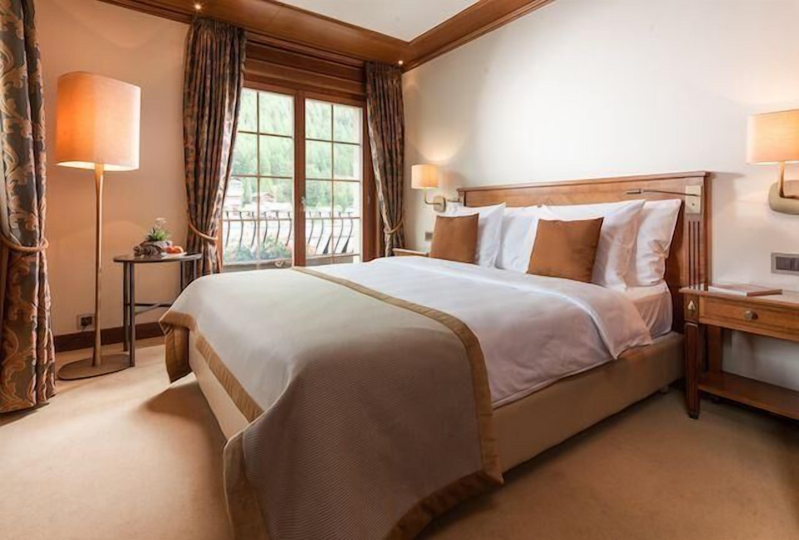A hotel room with a large bed, a nightstand, a lamp, and a view of the outdoors through a window.