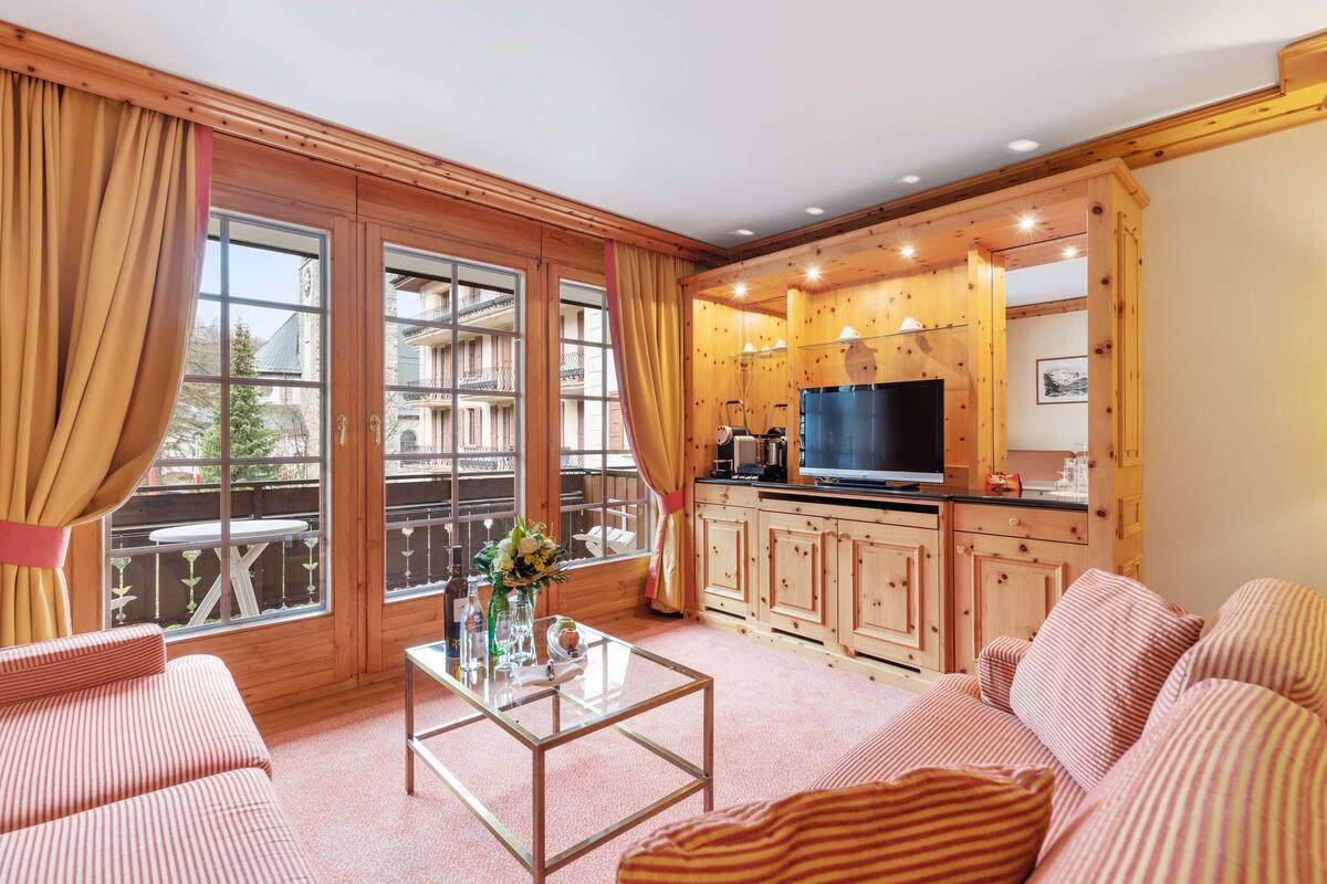 A cozy living room with a sofa, a coffee table, and a TV. There is a kitchen area with wooden cabinets and a large window.