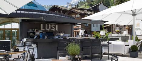 Un área de comedor al aire libre con mesas y sillas, una barra con rótulos de 'LUSI', y una vista de la montaña al fondo.
