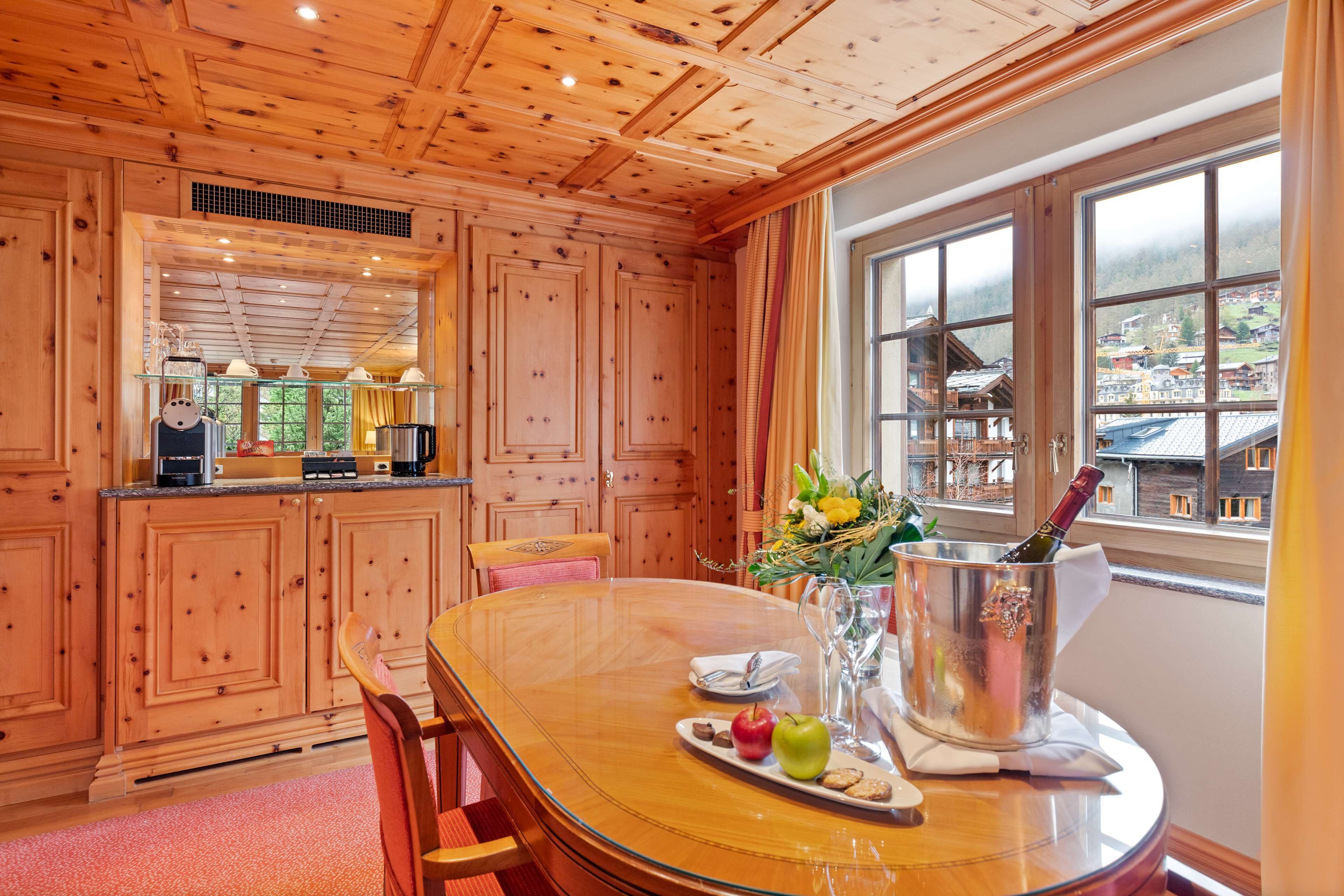 A wooden interior with a dining table set for two, a wine bottle in a bucket, and a view of a village through large windows.