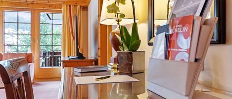 A hotel room with a wooden ceiling, a desk with a lamp, a plant, and a magazine rack.