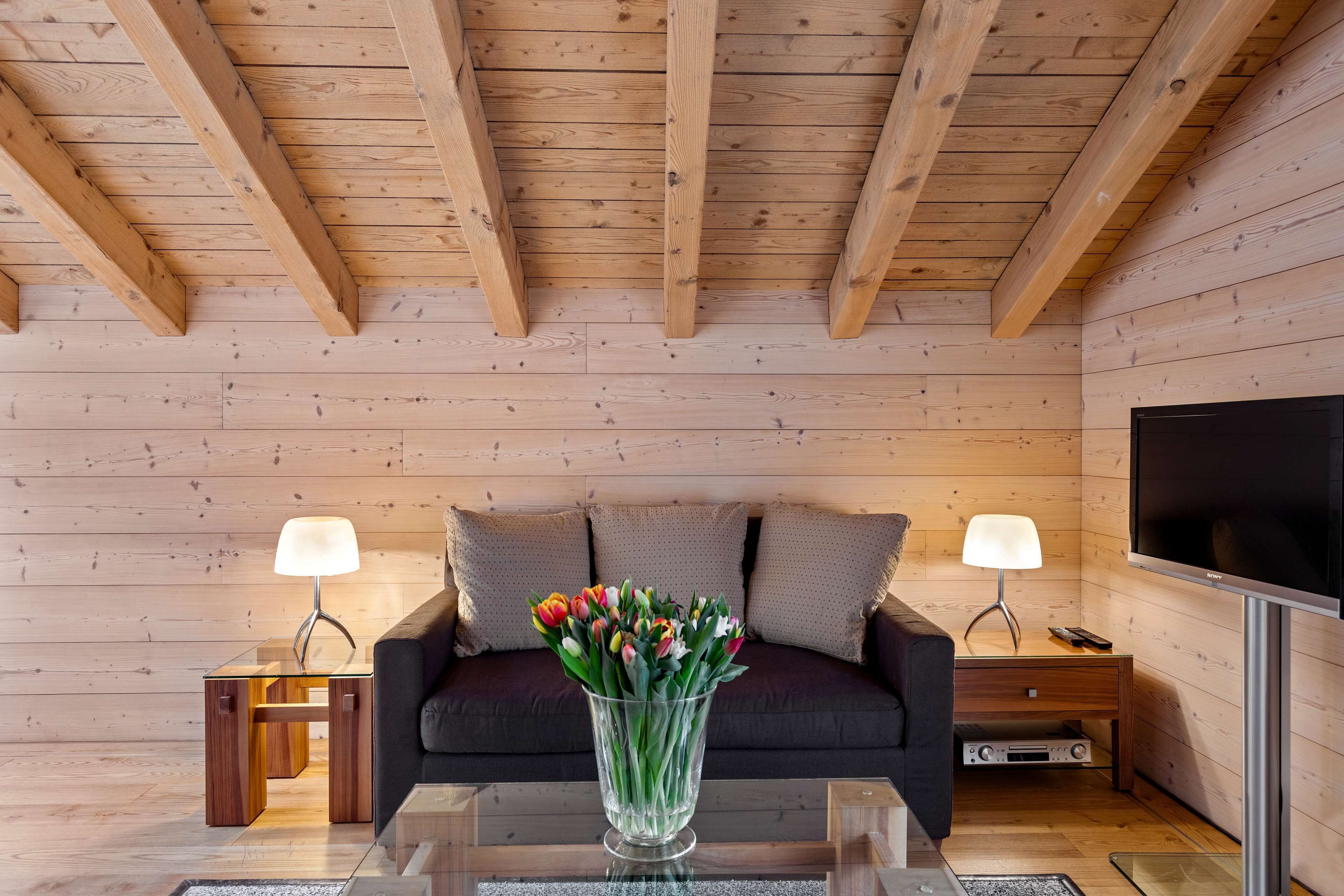 A wooden-paneled room with a sofa, two side tables with lamps, a TV, and a glass coffee table with a vase of flowers.