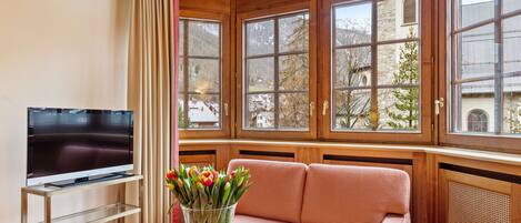 A room with a television on a stand, a sofa, a glass table with a vase of flowers, and large windows with a view of buildings and trees.