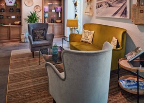Una sala de estar moderna con un sillón gris, un sofá color mostaza y una mesa de centro de vidrio. Hay una estantería con varios objetos y un cuadro en la pared.