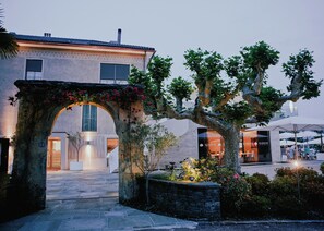 An outdoor area with a stone archway, a tree, and a building with a large window and a door. - Giardino Lago (Minusio)