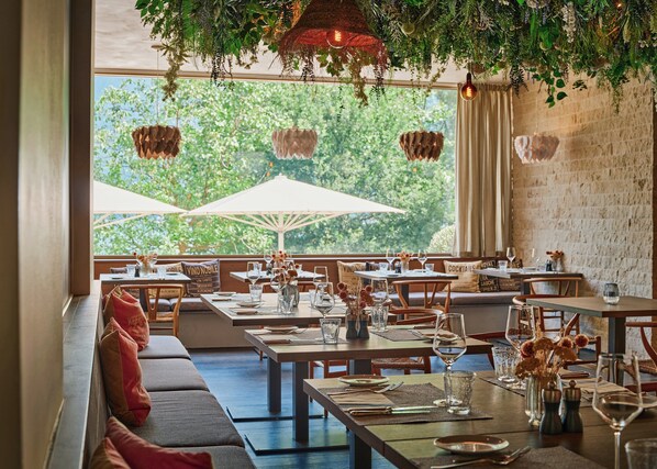 A restaurant with outdoor seating, tables set with wine glasses and cutlery, and hanging plants above. - Giardino Lago (Minusio)