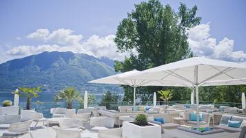 A terrace with white wicker furniture, umbrellas, and potted plants, overlooking a lake and mountains.