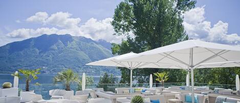 Une terrasse dotée de meubles en osier blanc, de parasols et de plantes en pot, offrant une vue sur un lac et des montagnes.