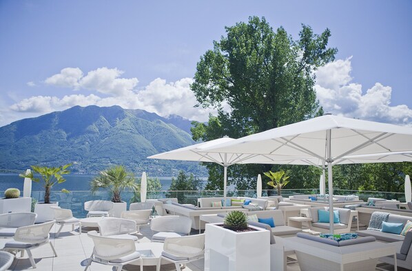 A terrace with white wicker furniture, umbrellas, and potted plants, overlooking a lake and mountains. - Giardino Lago (Minusio)
