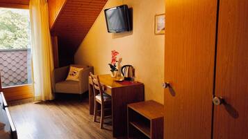 A cozy room with a wooden ceiling, a TV mounted on the wall, a sofa with a cushion, a dining table with chairs, and a small shelf.