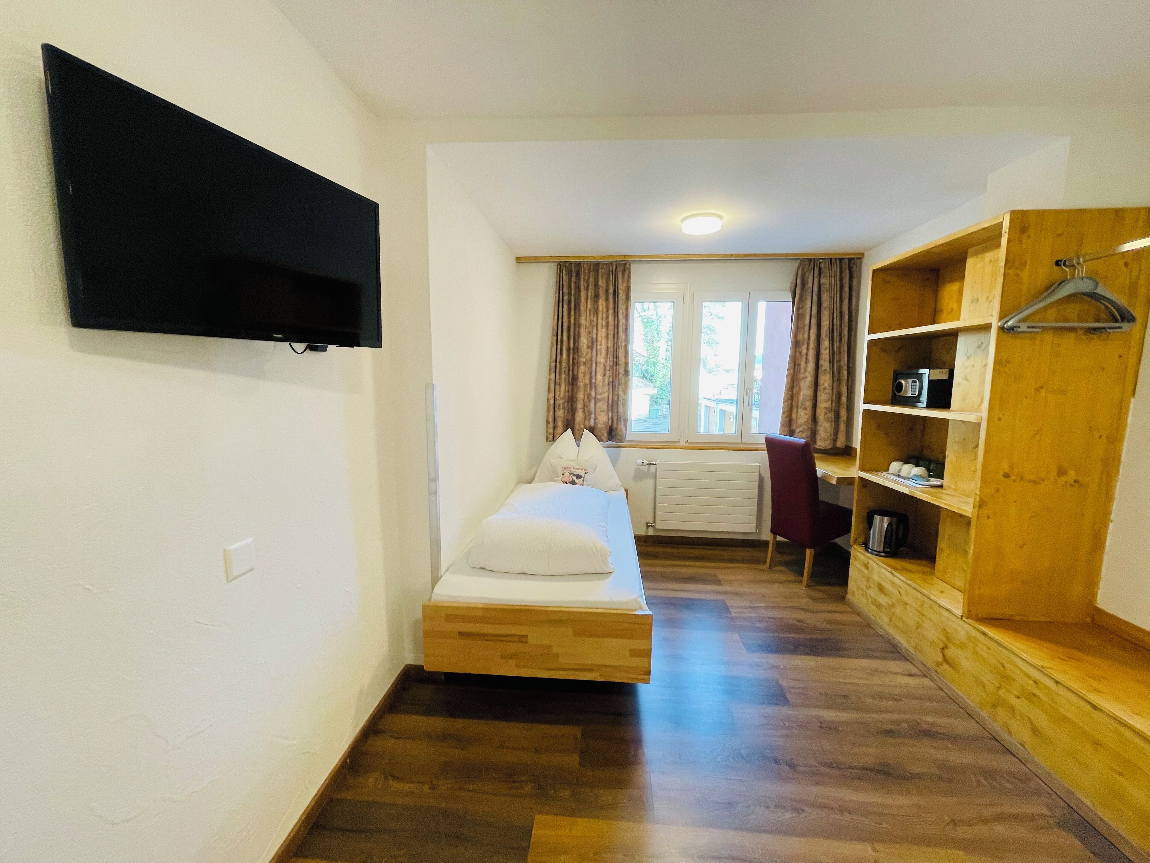 Ein kleiner, kompakter Raum mit einem Bett, einem an der Wand montierten Fernseher, einer Holzregaleinheit mit Kleiderstange und einem Fenster mit Vorhängen.