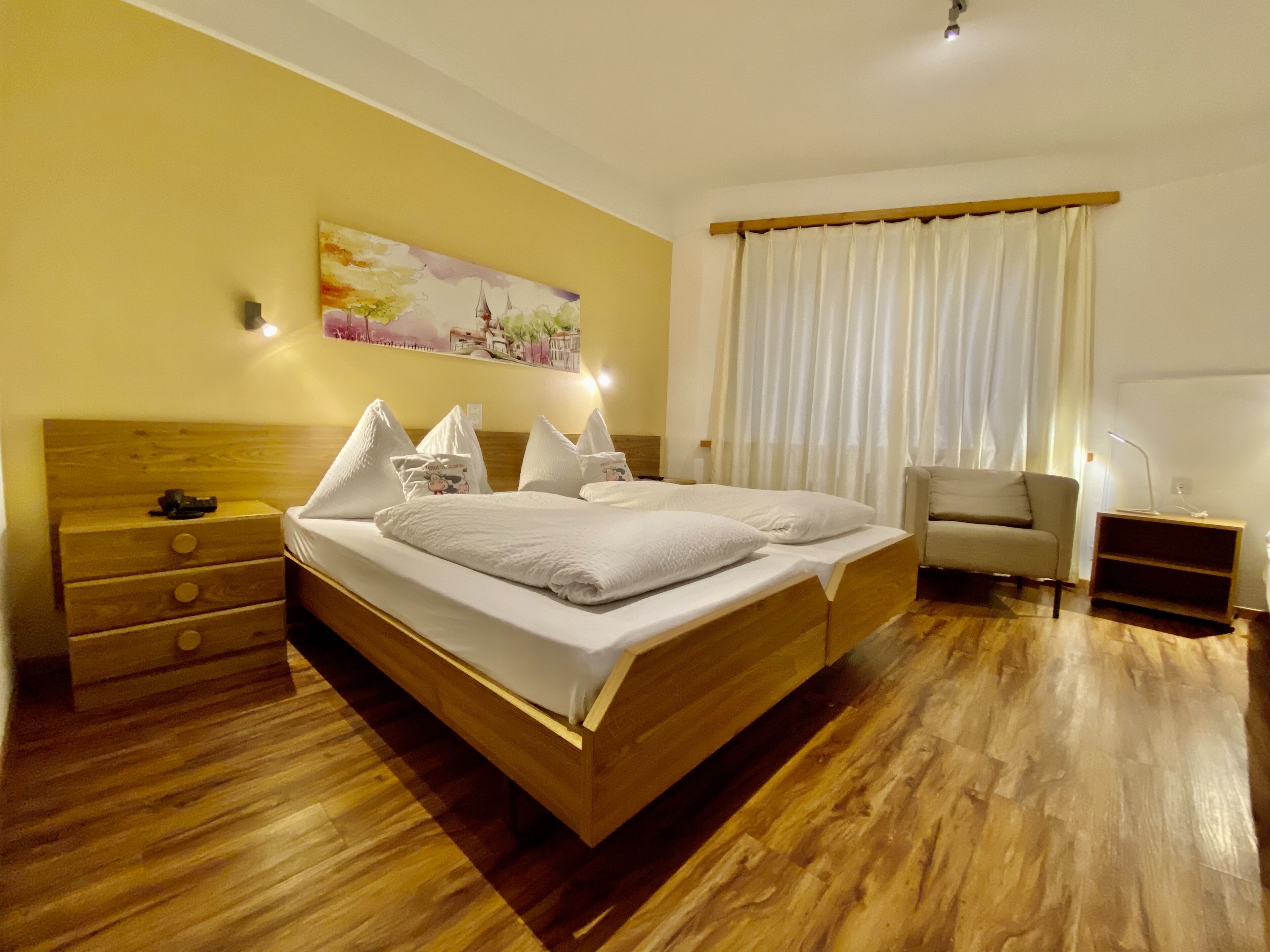 A hotel room with a wooden bed, a nightstand, a chair, and a painting on the wall.