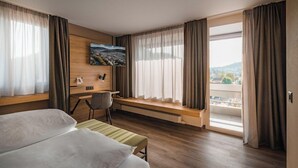 A hotel room with a bed, a desk, a chair, a window with curtains, and a painting on the wall. - Hotel Linde (Baden)