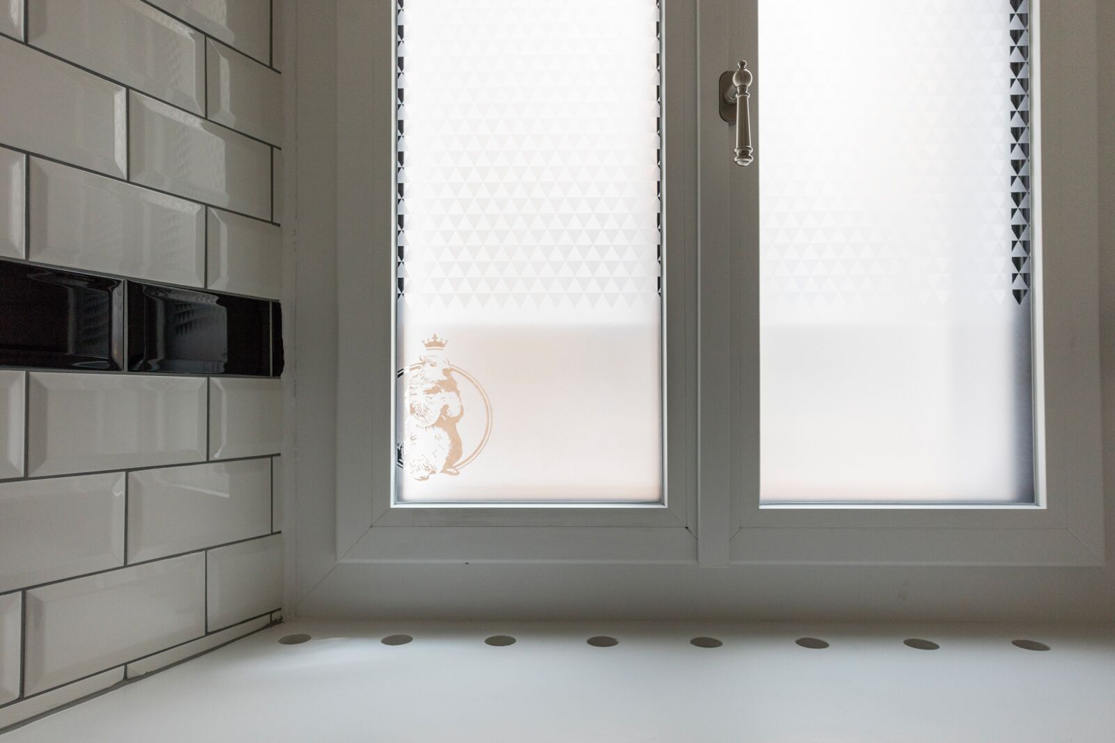 A modern bathroom with a white tiled wall, a double door with frosted glass panels, and a visible logo on the door.