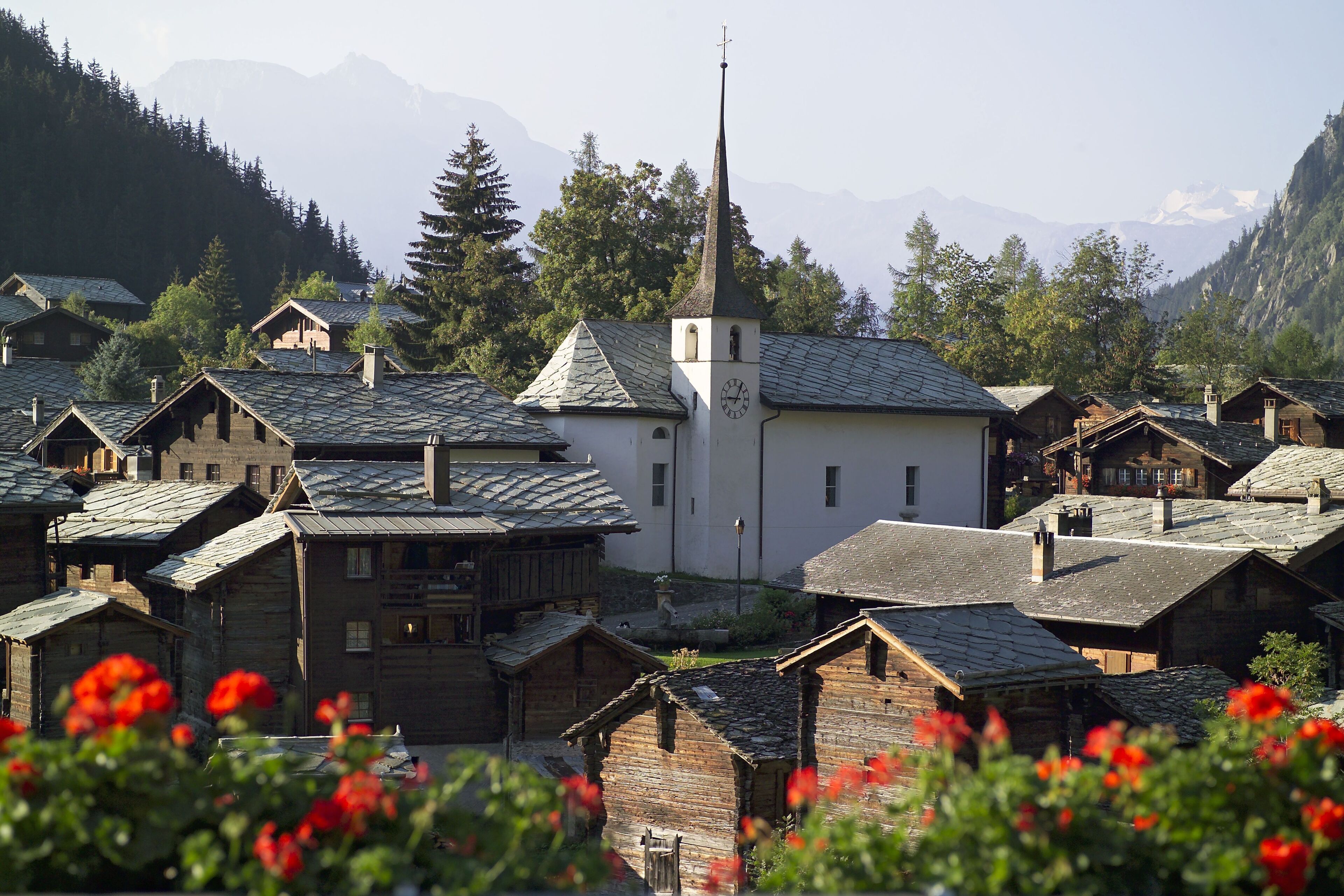 Un pueblo con casas de madera tradicionales, una iglesia con campanario y montañas al fondo.