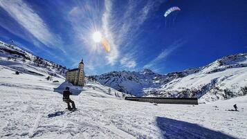 Paisaje nevado con un edificio, una persona practicando snowboard y un paracaídas en el cielo.
