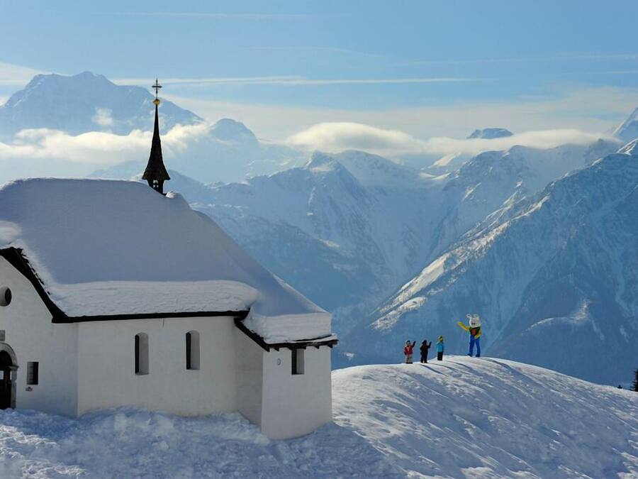 Una iglesia cubierta de nieve con una cruz en la parte superior, rodeada de montañas nevadas y un cielo despejado.